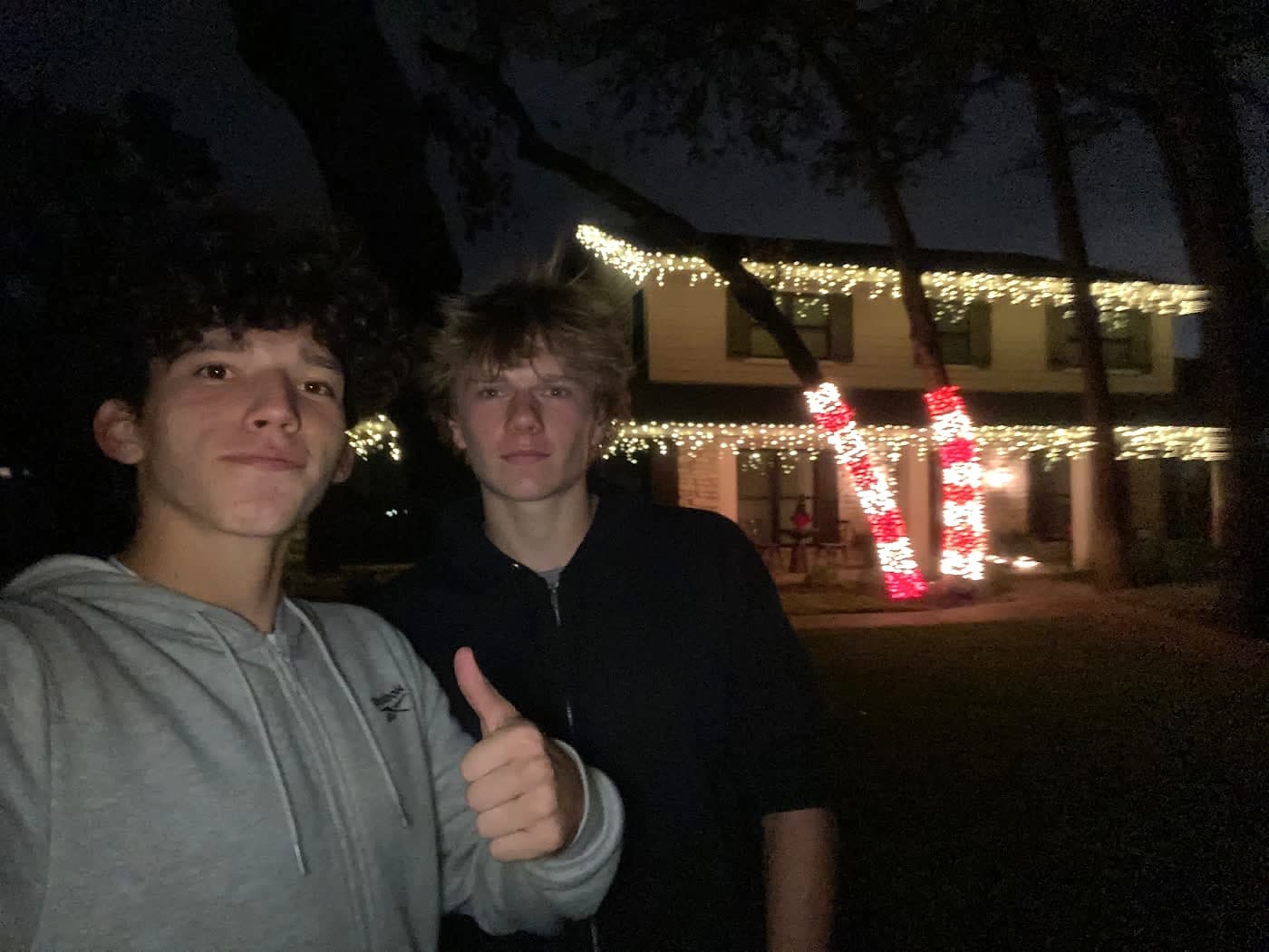 Graydon and Juandi standing in front of a two-story house they have just lit, with icicle lights along the roofline and candy-cane wrapped columns