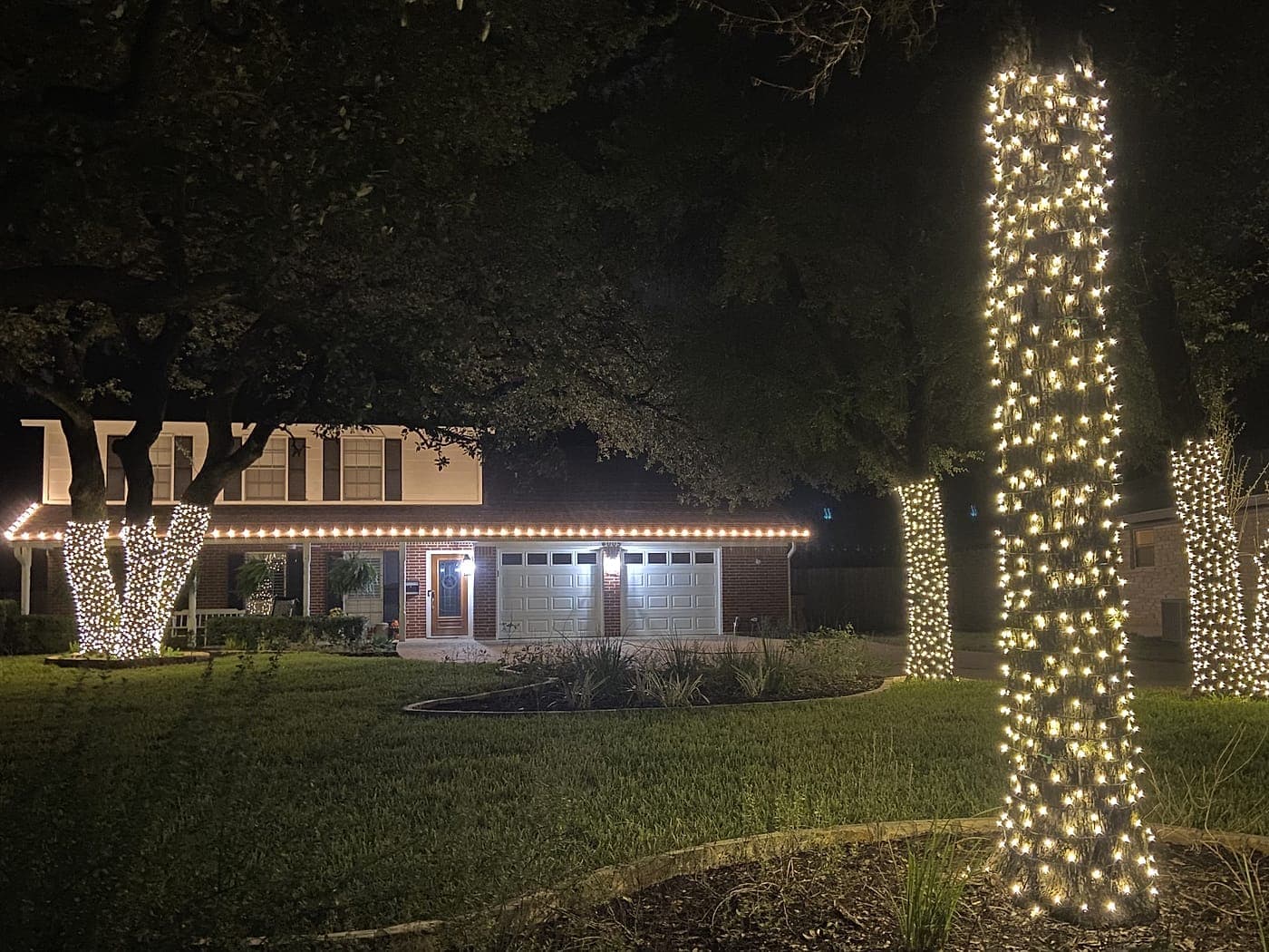 Three trees with trunks wrapped in warm white lights in front of a two-story home with a lit roofline